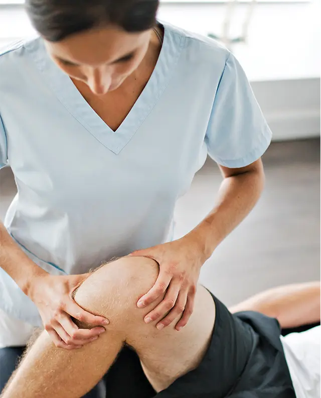 **Alt text:** A physical therapist in light blue scrubs gently examines and supports a patient’s knee during a therapy session at Katmai Eye and Vision Center, focusing on rehabilitation and improved mobility.